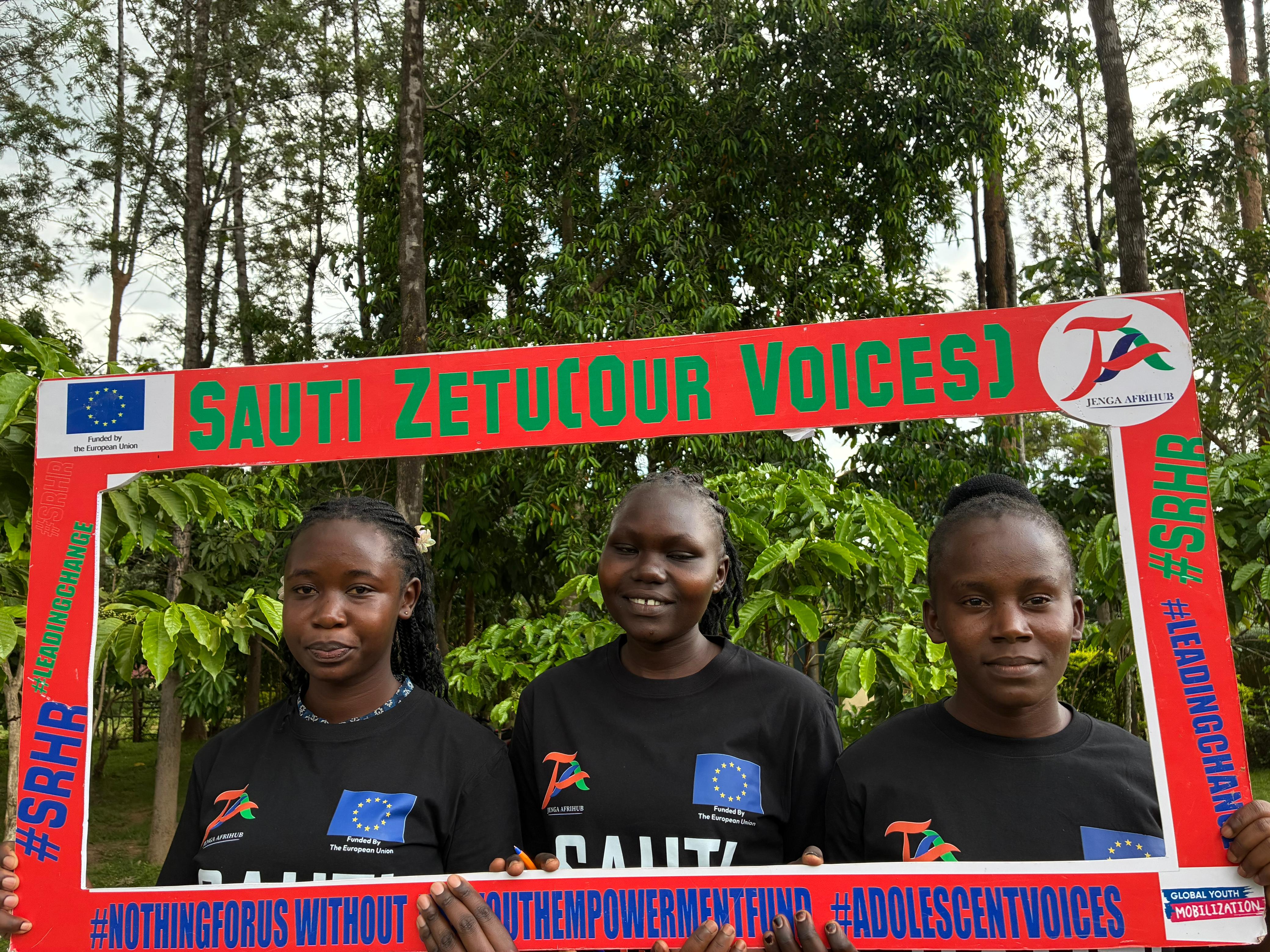 Three adolescent girls in Sauti Zetu shirts posing with the Sauti Zetu Our Voices photo frame